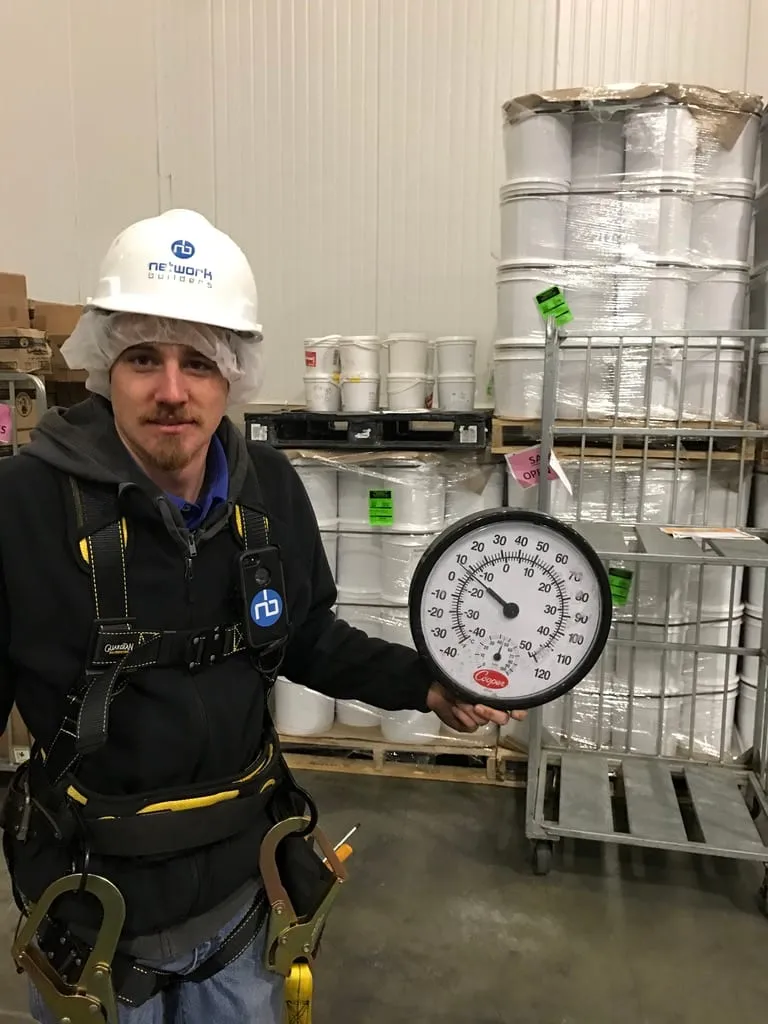 An NBIT engineer in cold-storage PPE and a safety harness inside a food-production warehouse, holding a temperature gauge